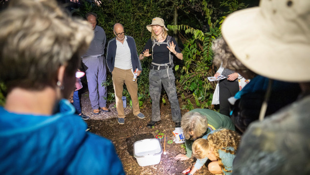 Een bodemdierengids met safarihoed neemt een groep mensen mee het arboretum in. De mensen luisteren, en sommige mensen zijn al bodemdieren aan het zoeken. Het is donker.